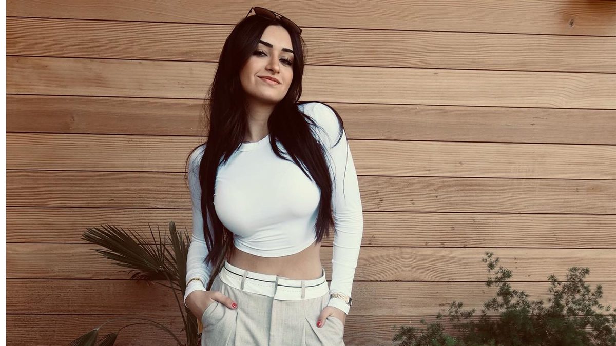 Christina Roki wearing white crop top and beige pants, standing on front of wood paneled wall